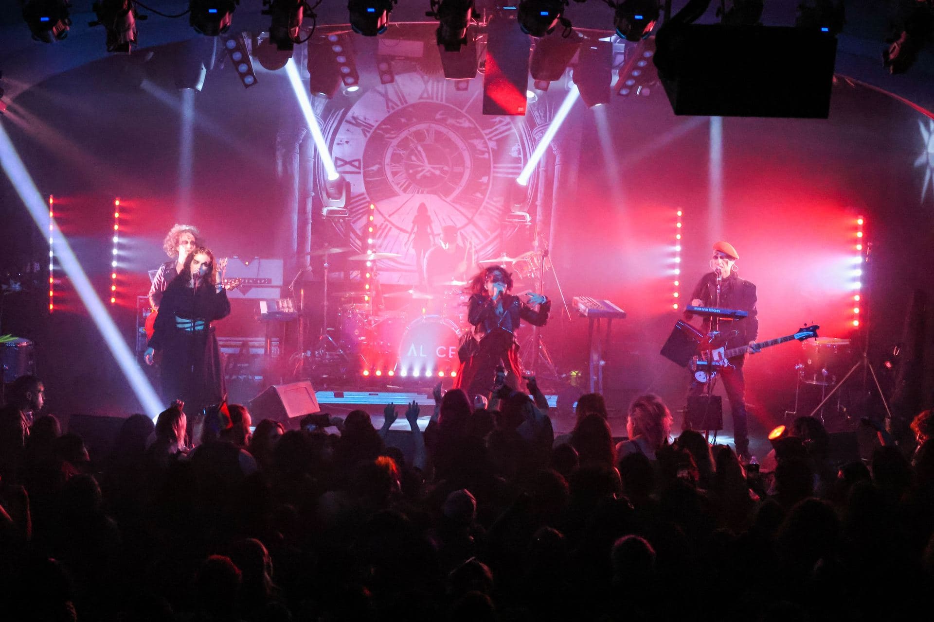 AL1CE performing to a packed room under red and white stage beams, the clock backdrop lit behind the band