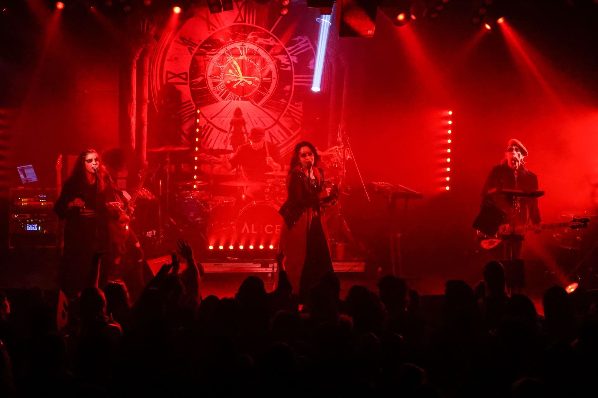The full AL1CE line-up in red stage light in front of the clock backdrop, crowd in silhouette