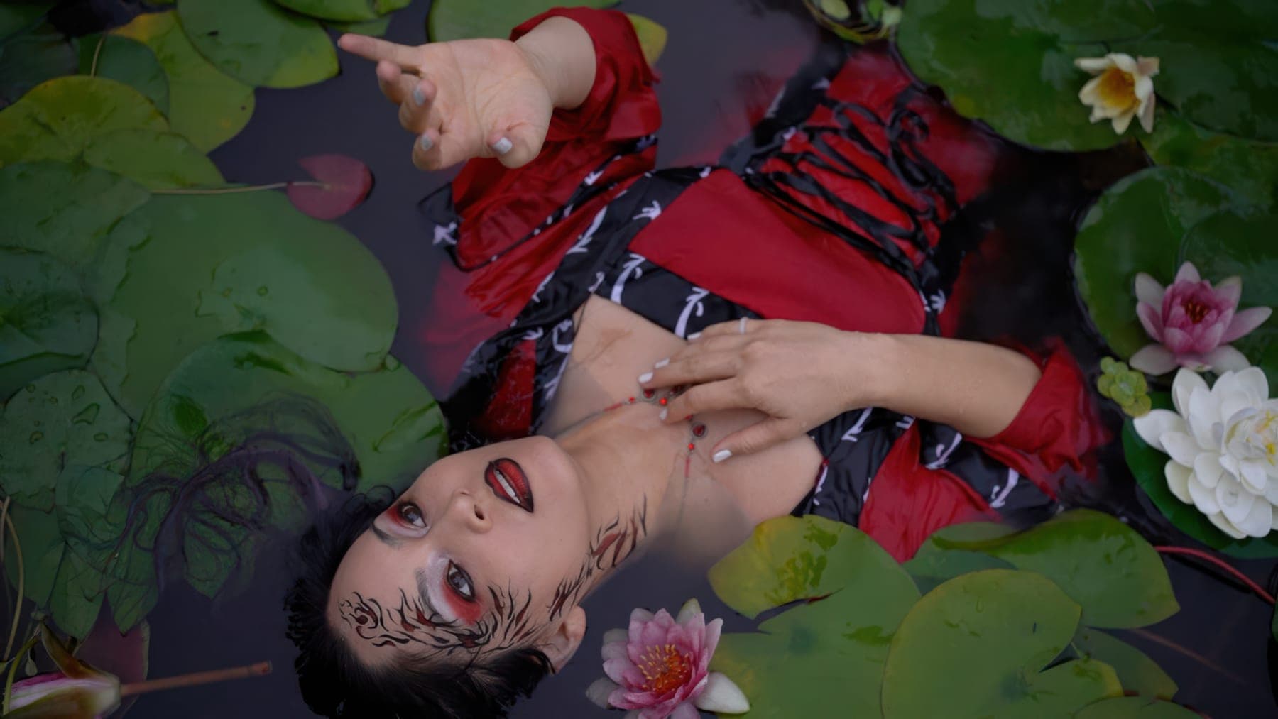 AL1CE vocalist floating among lily pads in ritual red makeup and costume